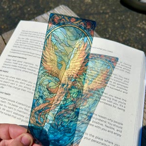 May include: A stained-glass style bookmark featuring a phoenix with outstretched wings in shades of gold, orange, and blue. The bookmark is held over an open book, showcasing the intricate design and vibrant colors. A unique and artistic accessory.