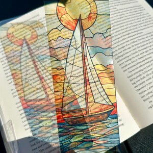 May include: A transparent bookmark featuring a sailboat design with a stained-glass effect. The image includes a sun, clouds, and water, with a book in the background. The bookmark is held up against the book.
