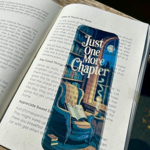 Puede incluir: Un marcapáginas con el texto "Just One More Chapter" sobre una ilustración de un rincón de lectura con una silla, una estantería y un libro abierto. El marcapáginas está en un libro abierto.