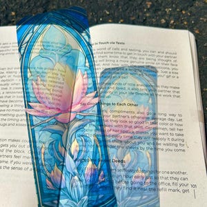 May include: A translucent bookmark with a stained-glass style design featuring a lotus flower in shades of blue, pink, and yellow. The bookmark is placed over an open book, with text visible in the background.