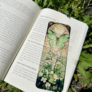 May include: A rectangular bookmark with a stained glass design featuring a green butterfly and white flowers. The bookmark is placed inside an open book with text visible on the pages.