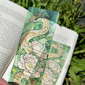 May include: A clear bookmark featuring a detailed illustration of a snake coiled around white and pink peonies. The design has a stained-glass effect with green and brown tones. The bookmark is placed over an open book.