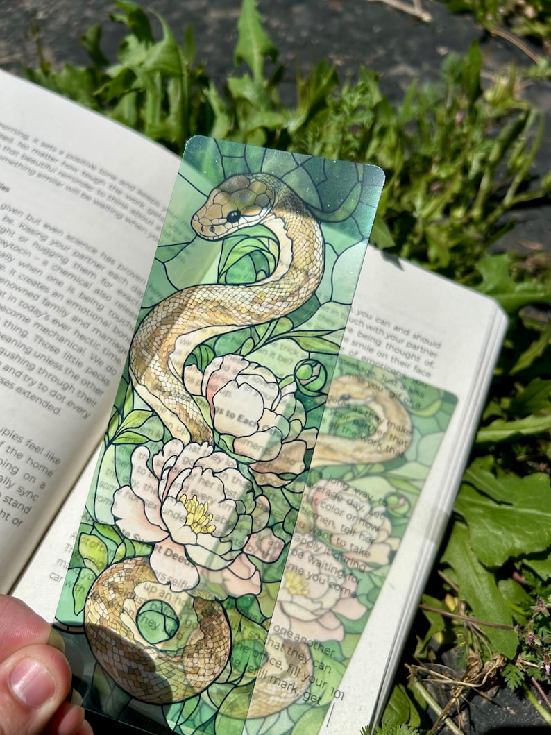 May include: A transparent bookmark with a snake and peony design, resembling stained glass. The snake is brown and beige, coiled around white and pink flowers. The bookmark is over an open book, with green and brown hues.