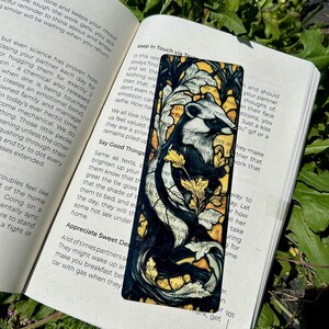 May include: A rectangular bookmark featuring a black and white illustration of a badger surrounded by yellow floral patterns and dark, swirling branches. The bookmark is placed on an open book with visible text, resting against green foliage.