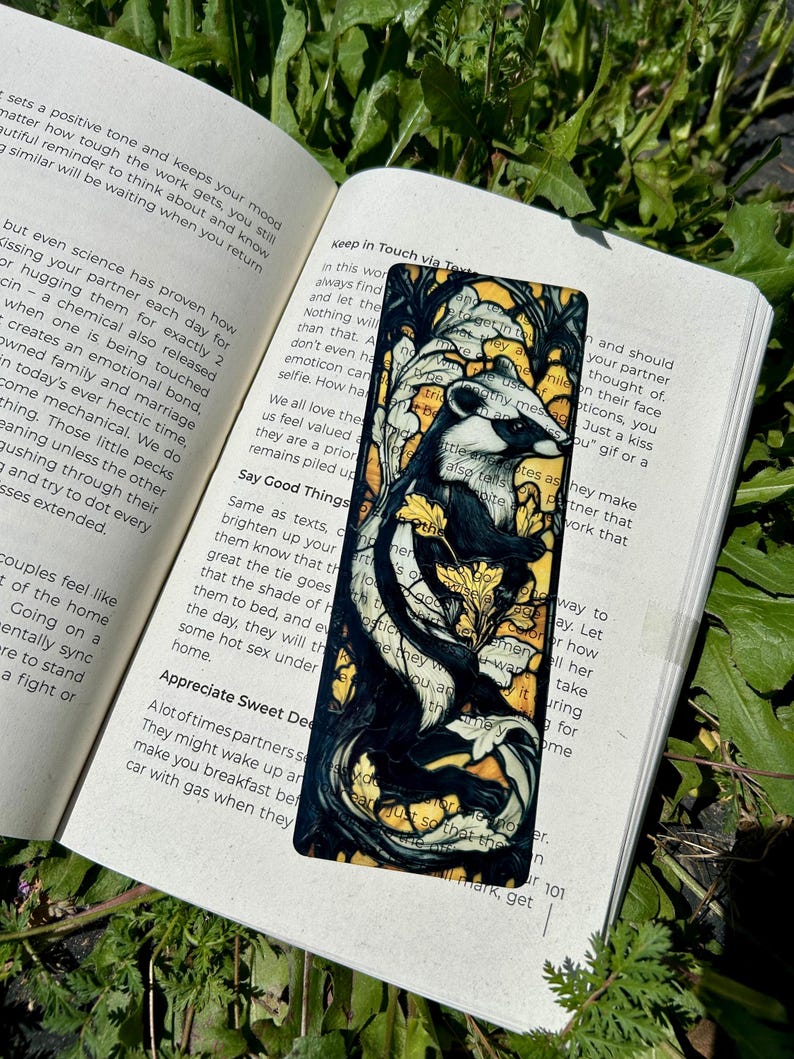 May include: A rectangular bookmark featuring a black and white illustration of a badger surrounded by yellow floral patterns and dark, swirling branches. The bookmark is placed on an open book with visible text, resting against green foliage.
