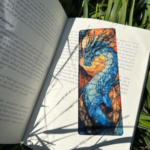以下が含まれることがあります： 青とオレンジのステンドグラスのブックマークで、細部までこだわったドラゴンが描かれています。ブックマークは長方形で、光沢のある仕上げになっています。