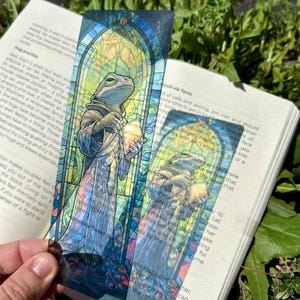 May include: A rectangular, semi-transparent bookmark featuring a stained-glass style illustration of a frog wearing a hooded robe and holding a book. The artwork uses vibrant blue, green, and yellow tones, framed by an arched architectural border.