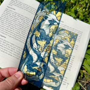 May include: Two decorative bookmarks featuring a badger illustration with a floral design in shades of blue, white, and gold. The bookmarks are held over an open book with text visible.