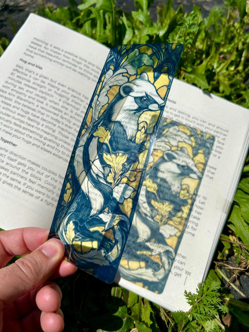 May include: A rectangular, semi-transparent bookmark featuring a detailed illustration of a ferret surrounded by stylized yellow and green foliage. The artwork uses dark blue outlines and a stained-glass aesthetic, set against a white book page background.