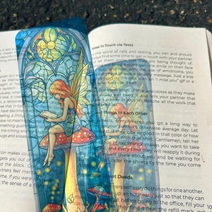 May include: A colorful bookmark featuring a fairy reading on a mushroom, set against a stained-glass window design. The bookmark has a transparent background and is placed over an open book. The fairy has red hair and yellow wings.