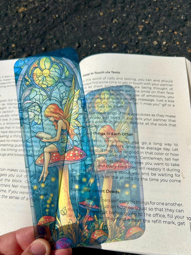 May include: A colorful bookmark featuring a fairy reading on a mushroom, set against a stained-glass window design. The bookmark has a transparent background and is placed over an open book. The fairy has red hair and yellow wings.