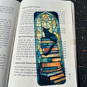 Puede incluir: Un marcap&aacute;ginas rectangular con un gato negro sobre una pila de libros, con un dise&ntilde;o de vidriera. El texto "Keep in Touch via Texts" es visible en la parte superior del marcap&aacute;ginas.