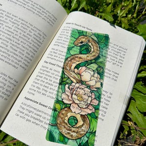 May include: A decorative bookmark with a detailed illustration of a snake, flowers, and a donut. The bookmark is placed in an open book, with text visible on the pages. The design has a whimsical and artistic style, with a green border.