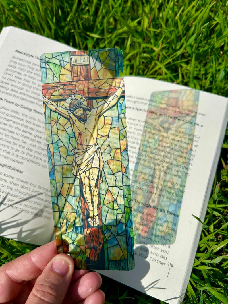 May include: A stained-glass style bookmark featuring a depiction of Jesus on the cross. The bookmark has a colorful mosaic design with the text "INRI" at the top. The bookmark is held over an open book on a grassy surface.