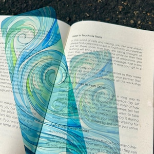 May include: A transparent, blue and teal-colored overlay with a swirling design is held over an open book. The book's pages display printed text. The overlay's design resembles water or a fluid pattern.