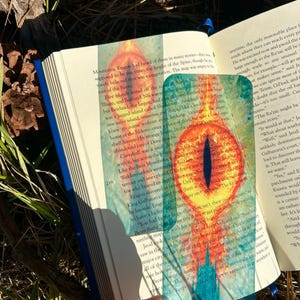 Könnte beinhalten: Transparenter Lesezeichen mit einer detaillierten Illustration eines Drachenauges in Orange, Gelb und Blau. Das Lesezeichen befindet sich in einem Buch, wobei Text sichtbar ist.