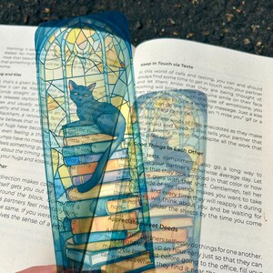 Puede incluir: Marcap&aacute;ginas transparente con un gato azul sobre una pila de libros. El marcap&aacute;ginas tiene un dise&ntilde;o de vidriera con colores azules, amarillos y blancos. El gato tiene una expresi&oacute;n seria.