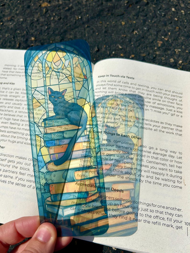 Puede incluir: Marcap&aacute;ginas transparente con un gato azul sobre una pila de libros. El marcap&aacute;ginas tiene un dise&ntilde;o de vidriera con colores azules, amarillos y blancos. El gato tiene una expresi&oacute;n seria.