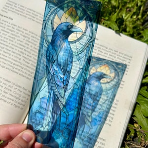 May include: A rectangular, translucent bookmark featuring a vibrant blue raven illustration set against a stained-glass style background with geometric patterns. The artwork displays deep shades of blue, teal, and black, with a circular motif behind the bird.