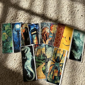May include: A selection of rectangular bookmarks featuring diverse designs. These include a stained-glass spider, a lion, a dinosaur, a smiling face, and a forest scene. The bookmarks are displayed on a textured, light brown surface.