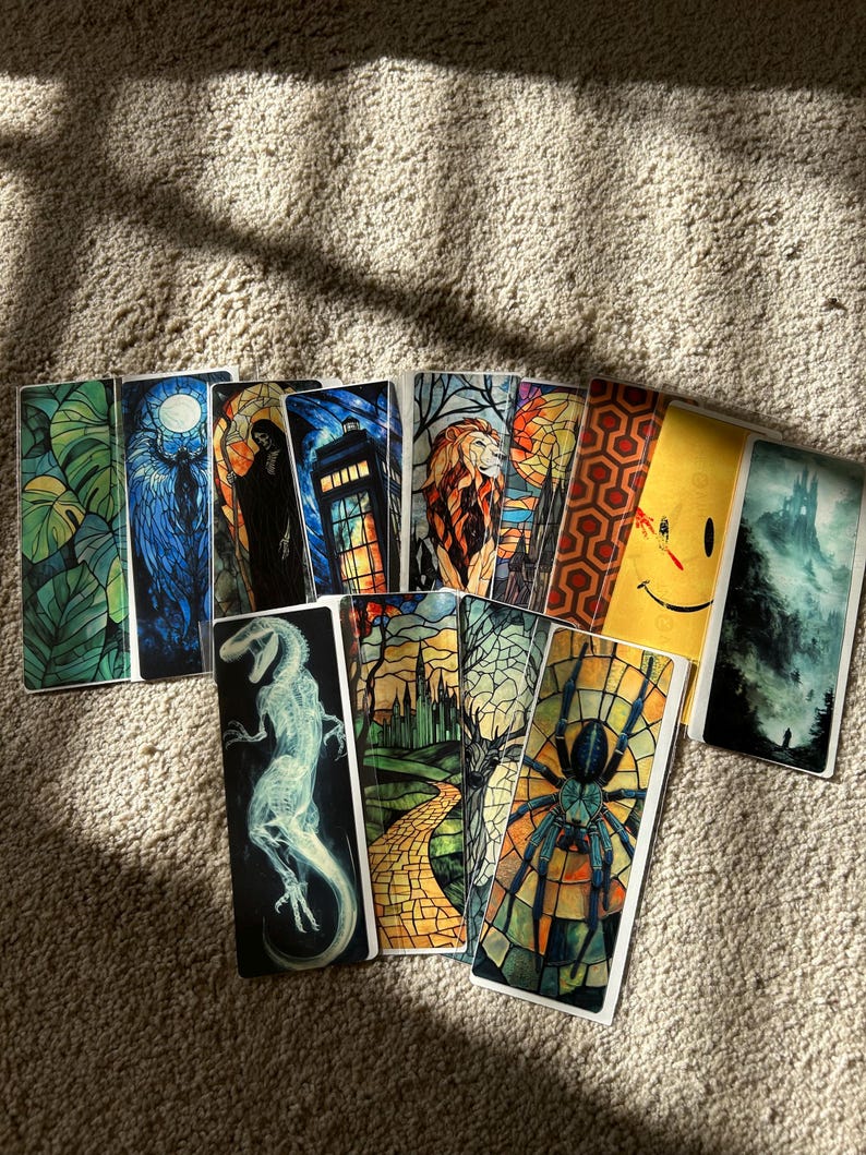 May include: A selection of rectangular bookmarks featuring diverse designs. These include a stained-glass spider, a lion, a dinosaur, a smiling face, and a forest scene. The bookmarks are displayed on a textured, light brown surface.