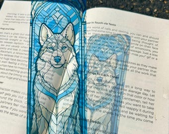 May include: A rectangular bookmark featuring a translucent blue stained glass design with a central illustration of a wolf. The wolf is depicted with intricate white and grey fur patterns, set against a geometric blue archway background.