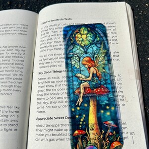 May include: A rectangular sticker featuring a fairy with red hair sitting on a mushroom. The fairy is reading a book, and the background is a stained-glass window design with colorful mushrooms.