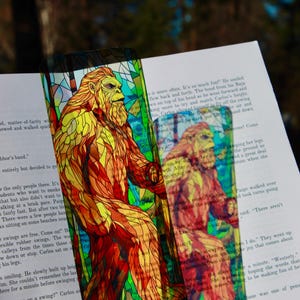 Può includere: Un segnalibro rettangolare con un'illustrazione in stile vetrata raffigurante una grande creatura bipede e pelosa nei toni dell'arancione, giallo e rosso. Il disegno è posto su uno sfondo geometrico scuro sopra la pagina di un libro aperto.