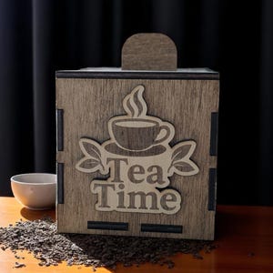 May include: Wooden tea box with a laser-cut design featuring a teacup, leaves, and the words "Tea Time." The box is brown, with a handle on top. A small white bowl and loose tea leaves are in the foreground.