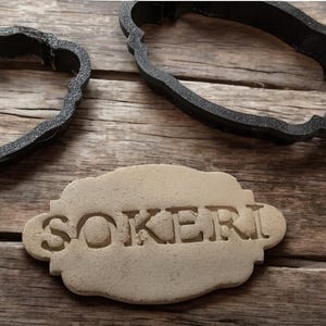 Może przedstawiać: Zbliżenie na beżowe ciasteczko z wytłoczonym napisem "SOKERI". W tle znajdują się dwa czarne foremki do ciasteczek.