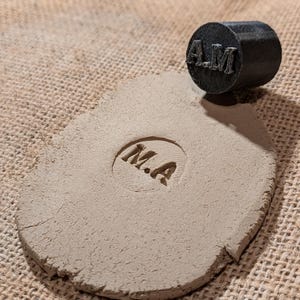 Könnte beinhalten: Nahaufnahme einer Tonscheibe mit den eingeprägten Buchstaben "M.A.". Ein schwarzer Gummistempel mit den Buchstaben "A.M." liegt auf einer Jutestoffoberfläche.