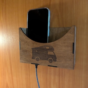 Pode incluir: Um suporte de telefone de madeira com um recorte para um telefone e um cabo de carregamento. O suporte tem um design de autocaravana e está montado numa parede de madeira.