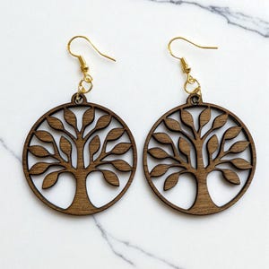 May include: A pair of wooden earrings featuring a tree of life design. The earrings are circular with a cutout tree motif and gold-colored ear wires. The wood has a warm brown tone, and the earrings are displayed against a white background.