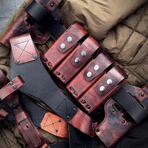 Op de afbeelding: Een verzameling handgemaakte, verweerde rode leren accessoires, waaronder holsters en riemen, met zwarte accenten en zilverkleurige klinknagels. De items zijn gerangschikt op een gewatteerde olijfgroene jas.