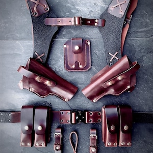 Puede incluir: Colección de accesorios de cuero marrón, que incluyen un arnés de pecho, fundas, cinturón y bolsas. Los artículos están dispuestos sobre una superficie oscura, mostrando la artesanía y el detalle del trabajo en cuero. El conjunto incluye herrajes metálicos.