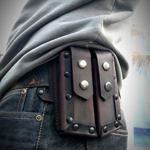 May include: A brown leather pouch with two compartments, secured with silver-colored snaps and rivets, attached to a belt. The pouch is worn on the back, over blue denim jeans.