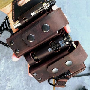 May include: A brown leather multi-tool holder with three compartments, each holding a metal pen or tool. The holder has a handle and is secured with metal press studs. The leather has a distressed finish.