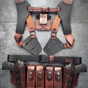 Puede incluir: Un conjunto de arnés y cinturón táctico de cuero en negro y marrón. El conjunto incluye un chaleco táctico con correas para los hombros, un cinturón con fundas y bolsas para cargadores. El cuero está envejecido, con costuras visibles y herrajes metálicos.