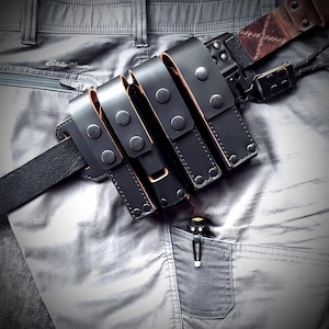 May include: A black leather multi-pouch belt attachment featuring four vertical compartments secured with metal snaps. The organizer is threaded onto a black leather belt, with a brown leather strap and metal hardware visible at the top right corner.