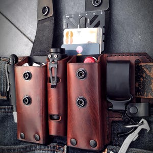 Könnte beinhalten: Ein brauner Leder-Gürtel-Organizer mit mehreren Taschen und einer schwarzen Gürtelschlaufe. Der Organizer ist an einer Jeans befestigt. Ein kleines silbernes Werkzeug in Form einer Axt hängt am Gürtel.