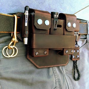Könnte beinhalten: Ein brauner Leder-Gürtel-Organizer mit mehreren Taschen und Fächern, an einem Gürtel befestigt. Enthält einen Stift, eine Taschenlampe und einen Schlüsselanhänger. Der Organizer ist an einer grauen Hose befestigt. Das Leder ist mit braunem Faden genäht und mit silbernen Nieten befestigt.