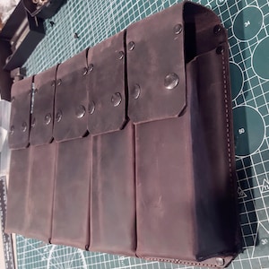 May include: A dark brown leather multi-pocket organizer with snap closures. The organizer has a rectangular shape with multiple compartments, each covered by a flap secured with a metal snap. The leather appears distressed, giving it a vintage look.