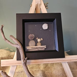 May include: A framed pebble art scene showing two figures under a starry night sky with a moon. The artwork is in a black frame, displayed on a small wooden easel, and accented with a piece of driftwood.
