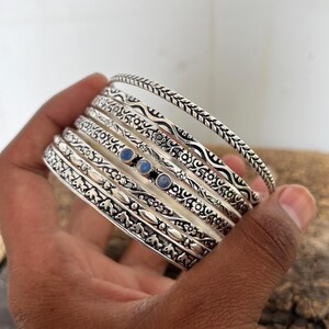 Könnte beinhalten: Eine Sammlung silberner Armreifen mit aufwendigen floralen und geometrischen Mustern. Einige Armreifen weisen kleine blaue Edelsteine auf. Die Armreifen werden in einer Hand gehalten, was die Vielfalt der Designs und Texturen zeigt.