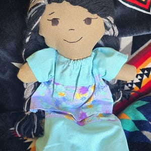 May include: A handmade doll with black and grey yarn hair, brown skin, and a blue and floral dress. The doll is sitting on a black and red patterned blanket.