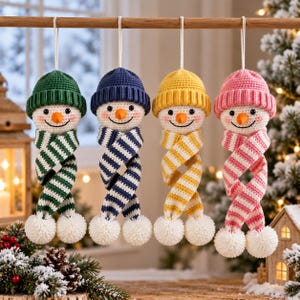 Op de afbeelding: Vier gehaakte sneeuwpop-ornamenten hangend aan een houten stok. Elke sneeuwpop draagt een effen muts in groen, blauw, geel of roze, en een bijpassende gestreepte sjaal met witte pompons aan de uiteinden. Ze hebben oranje wortelneuzen.