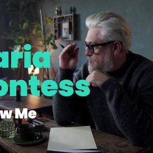 May include: The image displays the text "Maria Montess" in a teal gradient, with "Follow Me" underneath. A person wearing glasses and a beard is seated at a desk with a cup and a notepad. The setting is a room with indoor plants.
