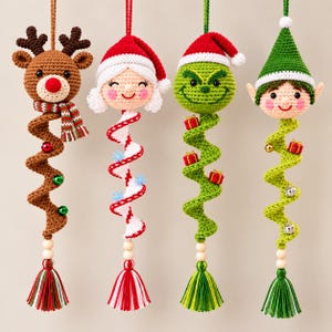 Könnte beinhalten: Vier hängende Häkelornamente mit einem Rentier, einer Weihnachtsmannfigur, einer grünen Kreatur und einem Elf. Jede Figur hat einen spiralförmigen Körper, Holzperlen und eine bunte Garnquaste am unteren Ende, verziert mit kleinen Geschenken und Glocken.