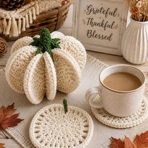 Pode incluir: Uma abóbora de crochê na cor creme com um caule verde escuro, um porta-copos redondo de crochê combinando e uma caneca de cerâmica cheia de café. Uma placa branca com o texto Grateful Thankful Blessed é visível ao fundo perto de flores secas.