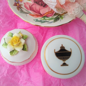 May include: Two white porcelain trinket boxes with gold trim. One box has a yellow rose with green leaves on the lid. The other box has a black and gold urn design on the lid.